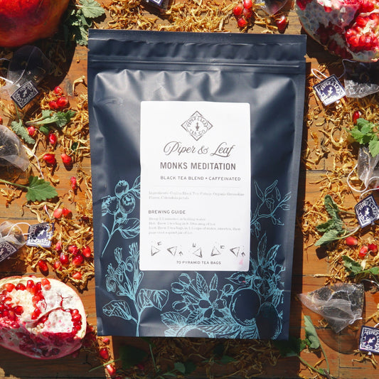 A package of Piper & Leaf Tea Co.'s Monks Meditation Bulk Sachets - 70ct Tea Bags, featuring Ceylon Black Tea, pomegranate, herbs, and catnip notes, rests on a wooden surface.