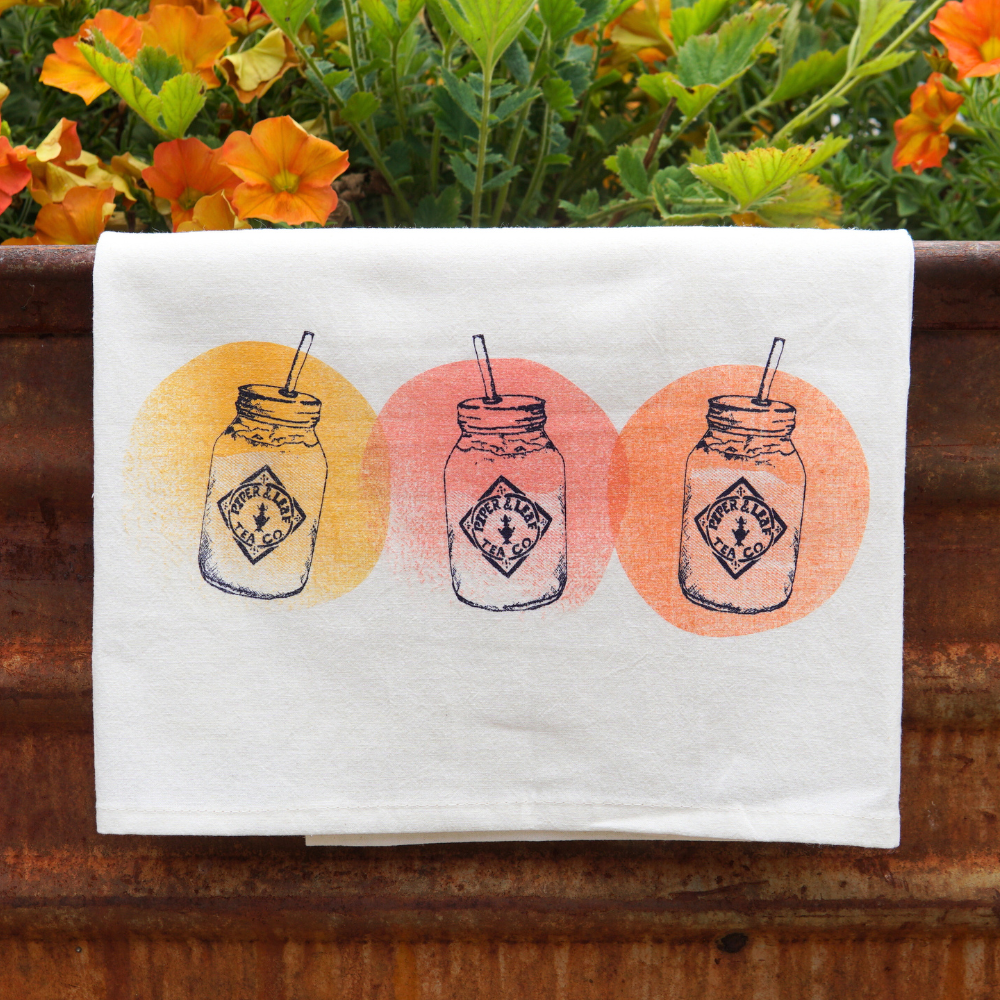 A white cloth with a print of three mason jars, each with a straw and a circular orange and yellow background, hangs over a rusted metal surface. Tea Towel- Mason Jars by Piper & Leaf Tea Co., featuring vibrant colors, complement the orange flowers and green leaves in the background.