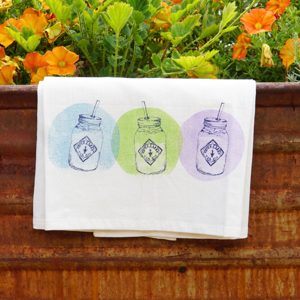 A lively screen-printed Tea Towel- Mason Jars by Piper & Leaf Tea Co., featuring three images of mason jars with straws, each surrounded by vibrantly colored circles (yellow, green, blue), hangs against a rusty metal surface with orange flowers in the background. Perfect for adding a pop of color to your kitchen accessories collection.