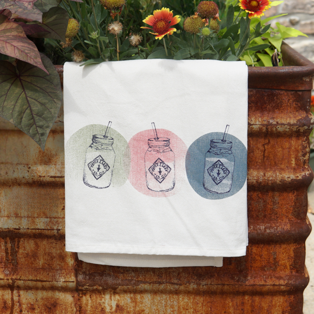 A white Piper & Leaf Tea Co. Tea Towel- Mason Jars with vibrant colors and mason jar prints in green, pink, and blue circles draped over a rusty barrel planter with flowers.