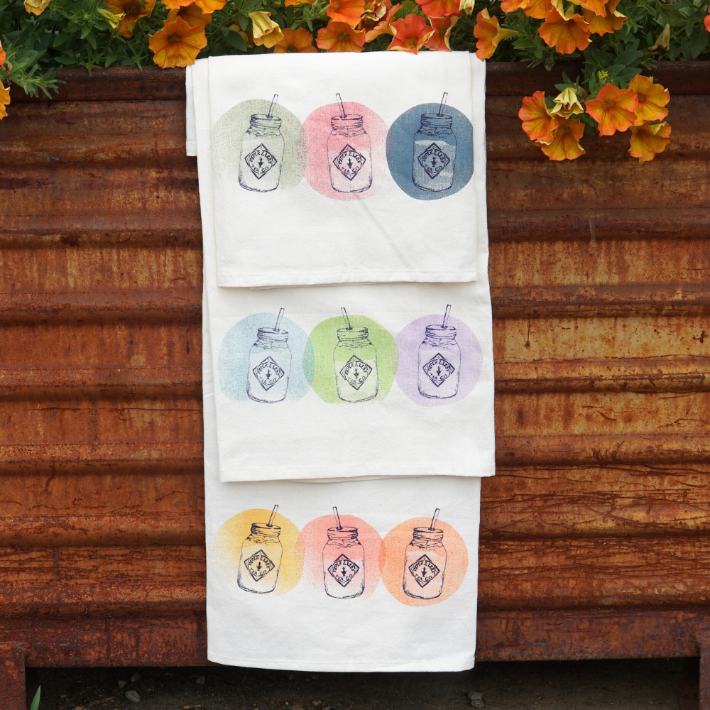 Three lively Tea Towel- Mason Jars from Piper & Leaf Tea Co. in vibrant colors are hanging on a rusted metal surface, with orange flowers adding a touch of charm in the background—perfect kitchen accessories to brighten up any space.