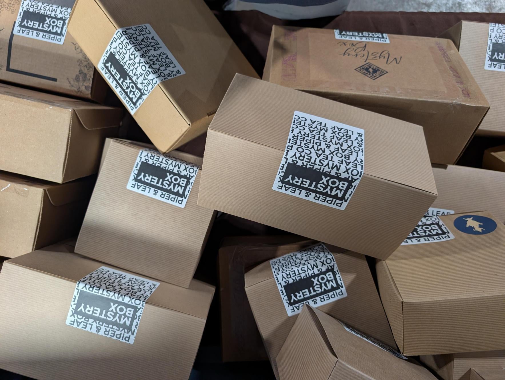 A stack of cardboard boxes, each labeled “Piper and Leaf Tea Co. Mystery Box,” hints at a unique tea gift experience from Piper and Leaf Tea Co.