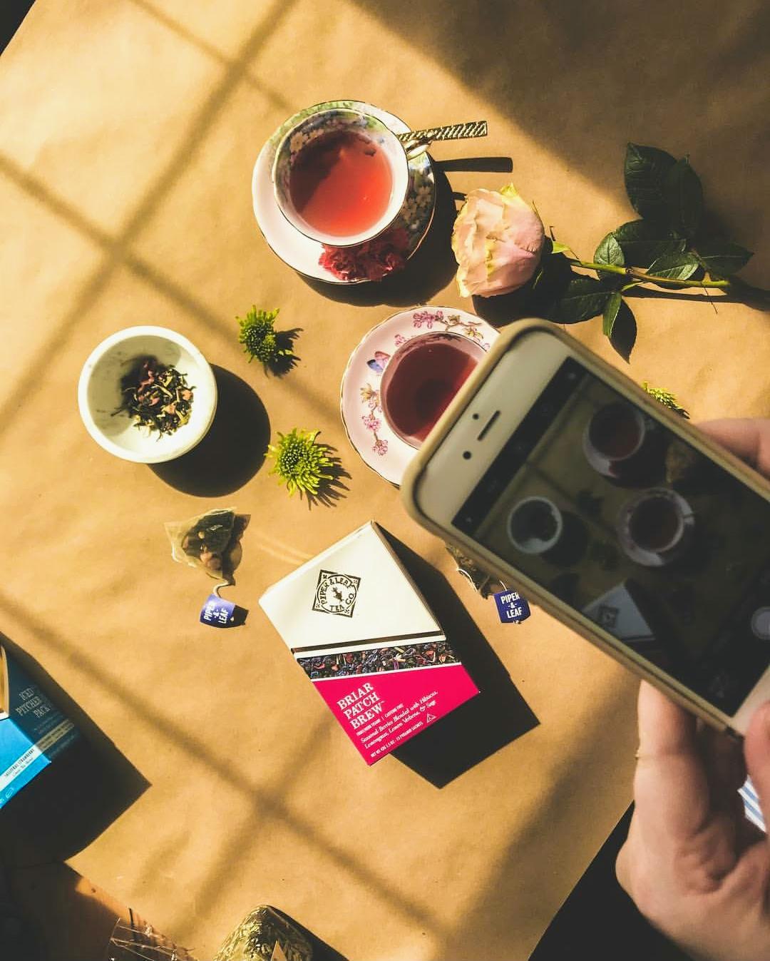 A person's hand holding a smartphone capturing a photo of a Springtime Afternoon Tea Party with cups, Piper and Leaf hot tea, and Mini Desserts on a sunlit table.