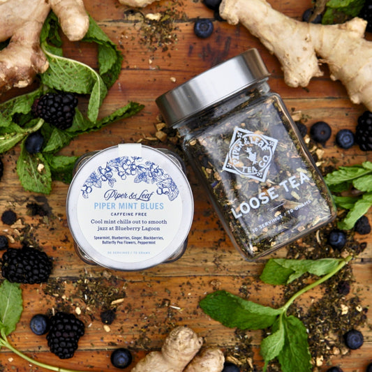 Two jars of Piper & Leaf Tea Co.'s "Piper Mint Blues," a loose leaf tea with 30 servings, are displayed on wood with ginger, mint, blackberries, and tea leaves. Savor the refreshing caffeine-free mint blend called "Blueberry Lagoon.