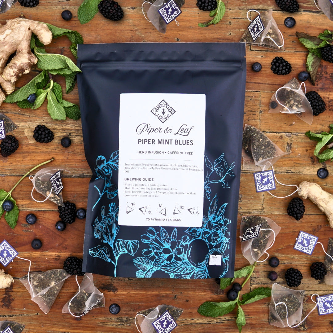 A bag of Piper & Leaf Tea Co. Piper Mint Blues Bulk Sachets - 70ct sits on a wooden surface, surrounded by ginger, fresh mint leaves, blackberries, blueberries, and scattered tea bags.