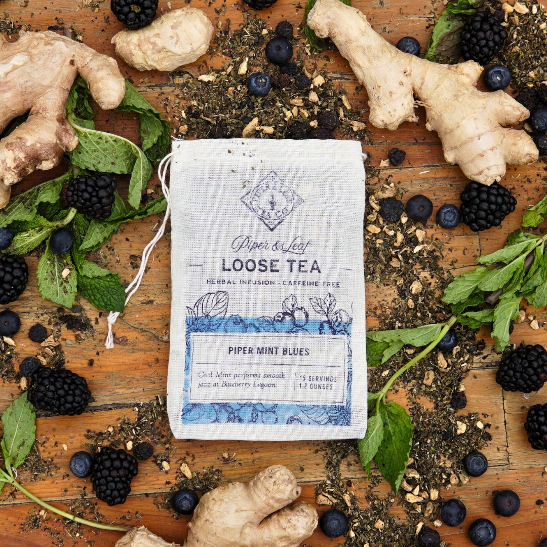 A Piper & Leaf Tea Co. muslin bag of "Piper Mint Blues" loose leaf tea, caffeine-free, displayed with ginger roots, blackberries, blueberries, mint leaves, and dried tea leaves on a wooden surface. Contains 15 servings.