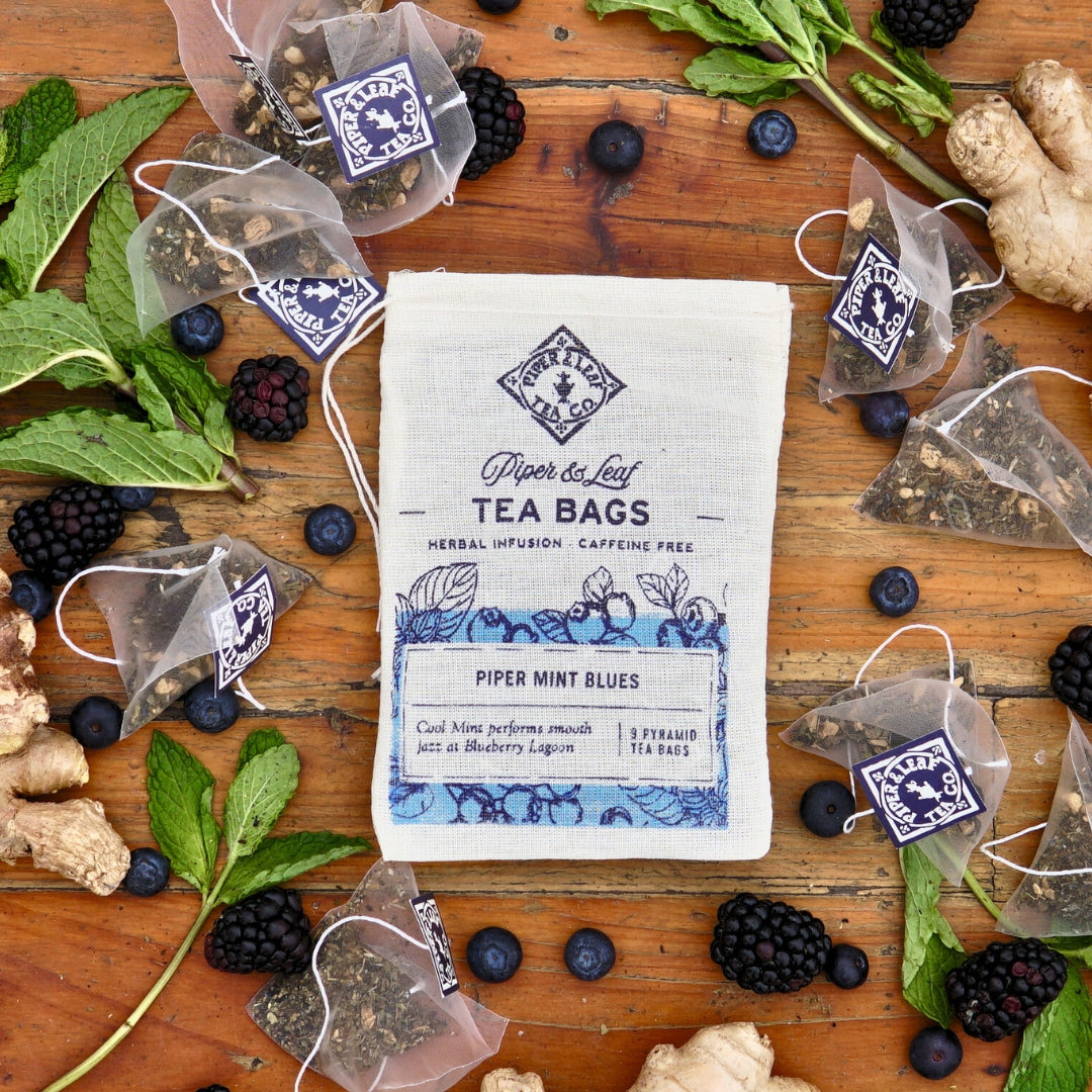 Piper & Leaf Tea Co.'s "Piper Mint Blues 9ct Tea Bags in Muslin" are displayed with fresh mint, juicy berries, blueberries, and ginger on rustic wood. This vibrant scene invites you to enjoy a refreshing mint tea experience that's naturally caffeine-free.