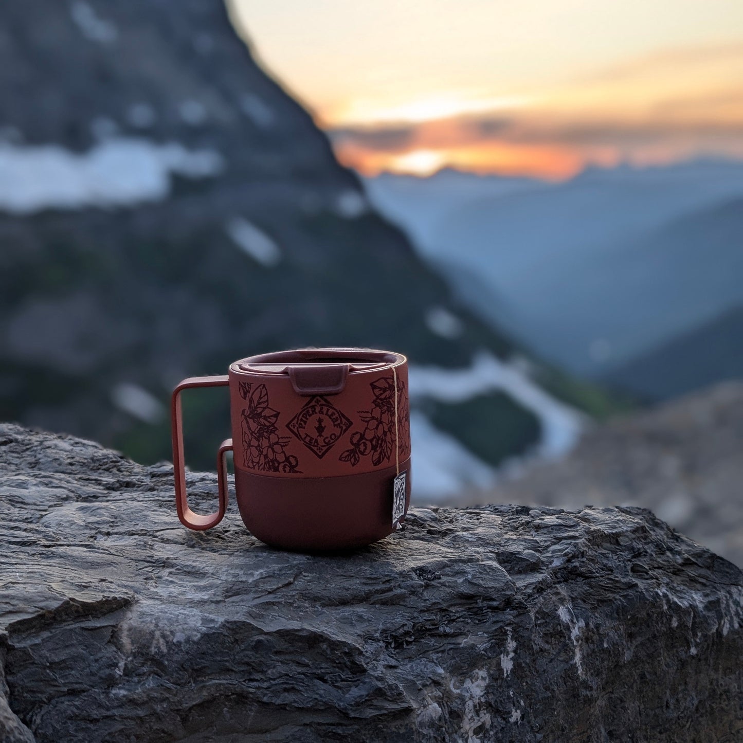 The P&L 14oz Rise Mug from Klean Kanteen by Piper & Leaf Tea Co. sits on a rocky surface, featuring red BPA-free design, with mountains and a sunset in the background.