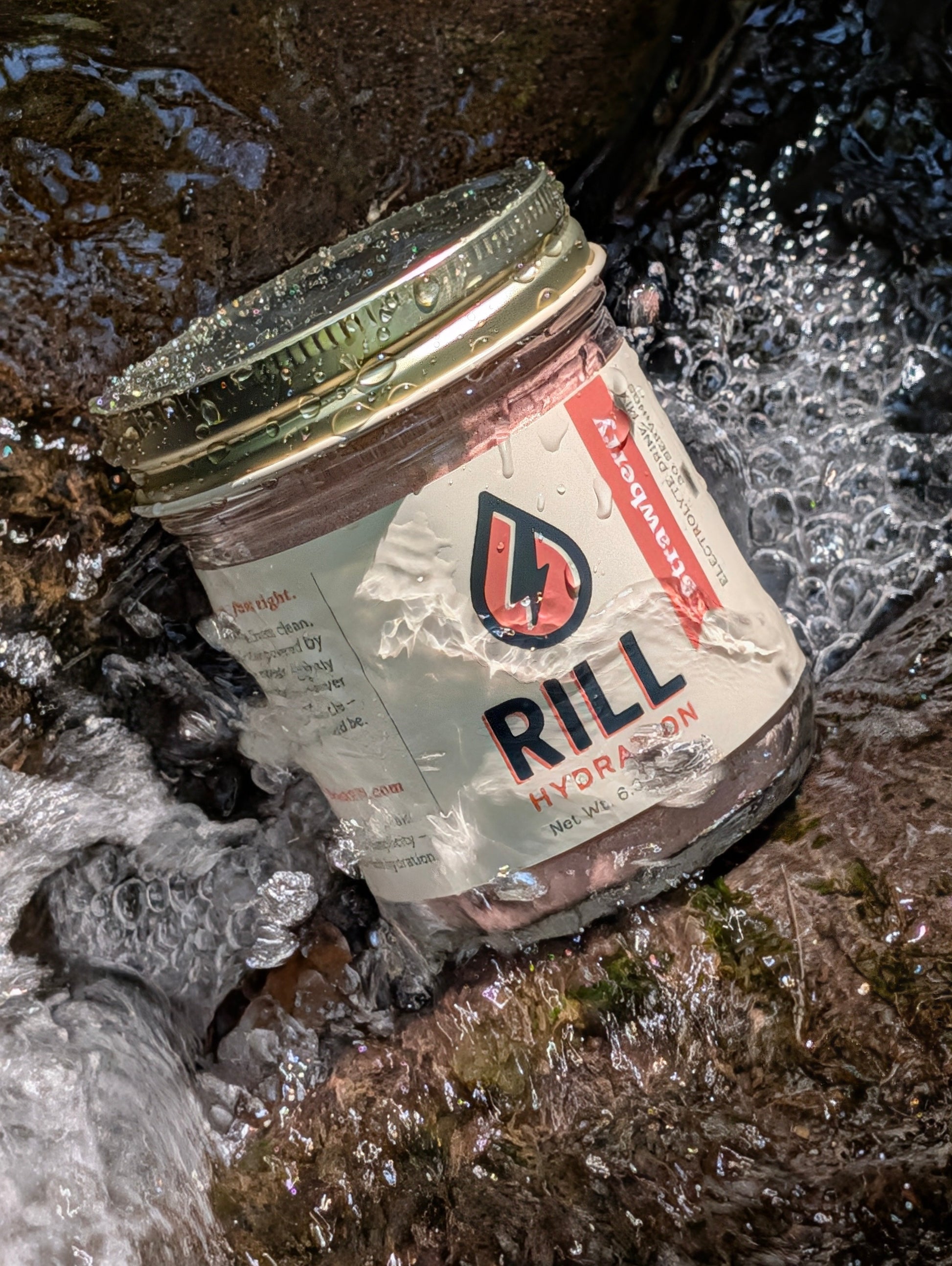 A "Strawberry Rill - Clean Hydration Drink Mix, 30 Serving Bulk Jar" from Rill Hydration rests partially submerged in flowing water, with droplets on its lid and label.