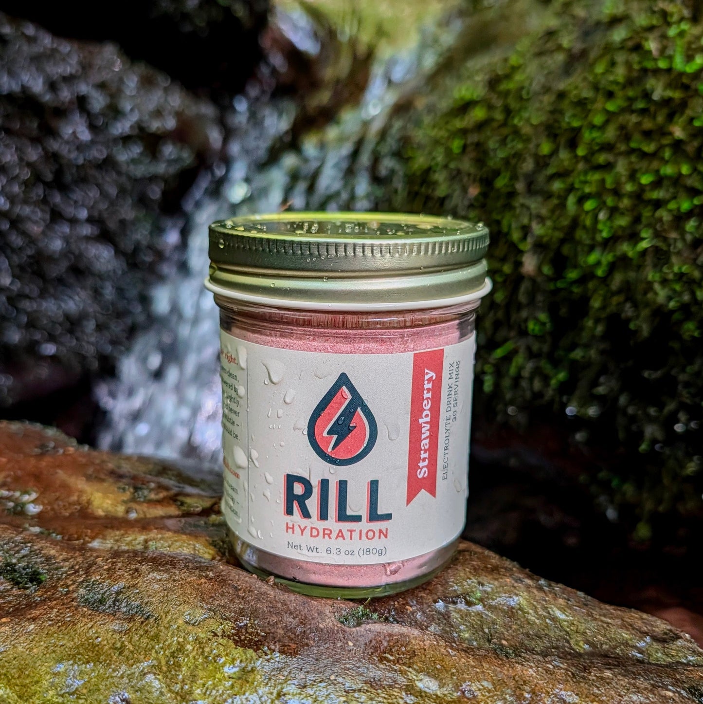 A Strawberry Rill - Clean Hydration Drink Mix, 30 Serving Bulk Jar by Rill Hydration sits on a wet rock amid moss and water droplets, showcasing this real fruit hydration powder.