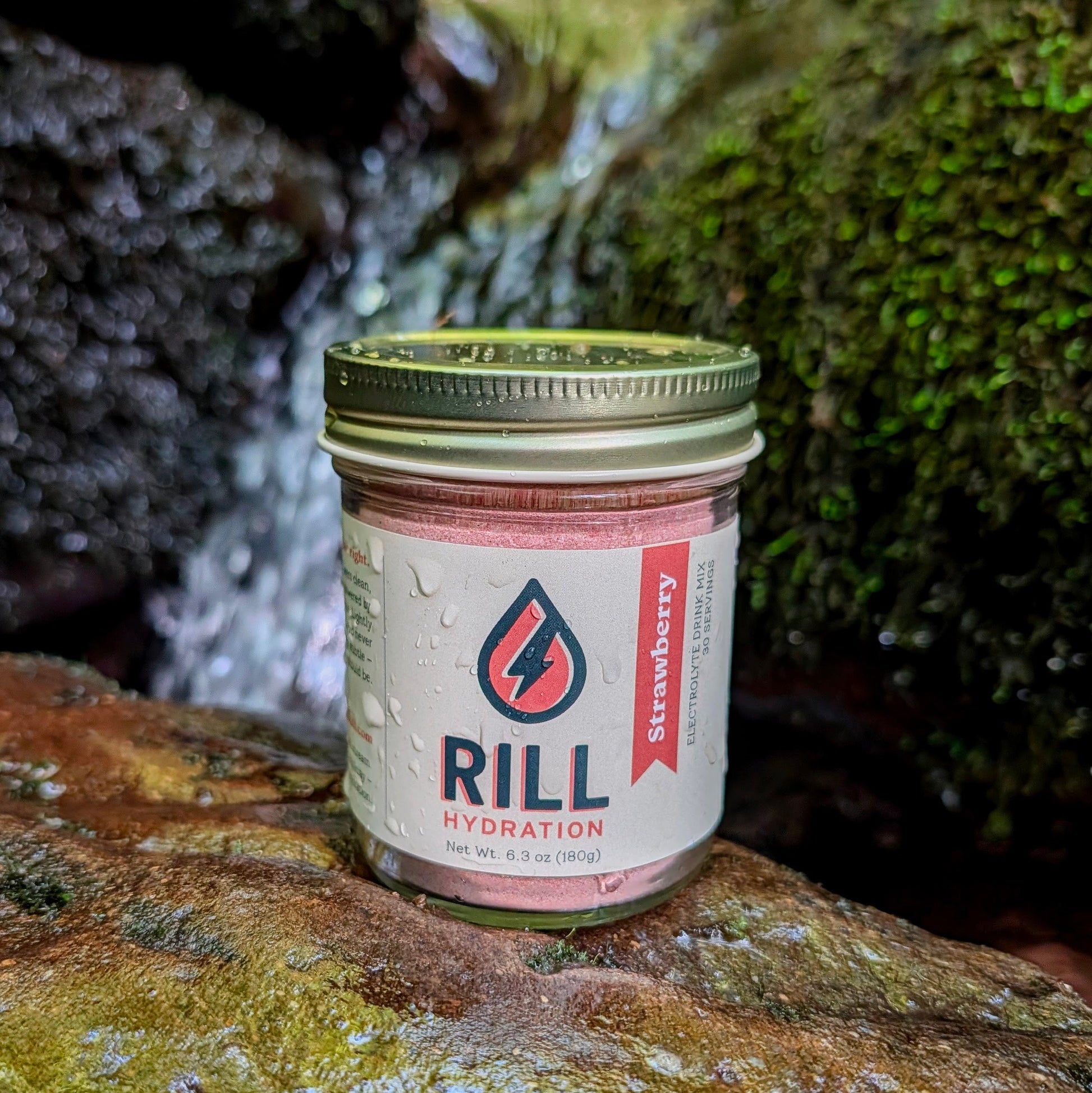 A Strawberry Rill - Clean Hydration Drink Mix, 30 Serving Bulk Jar by Rill Hydration sits on a wet rock amid moss and water droplets, showcasing this real fruit hydration powder.
