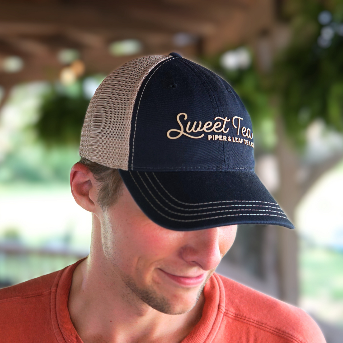 A man wearing a P&L Sweet Tea Ball Cap emblazoned with "Sweet Tea" and "Piper & Leaf Tea Co." looks down while clad in an orange shirt. The background is blurred, showcasing some greenery, reminiscent of the South's iconic beverage enjoyed on a warm day.