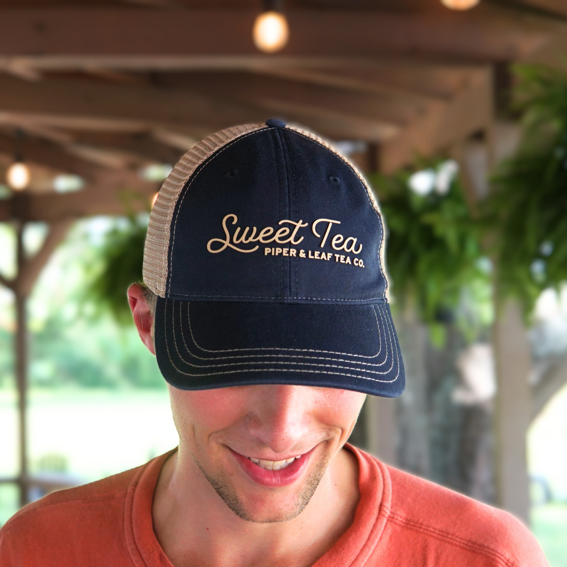 A man wearing a P&L Sweet Tea Ball Cap with the words "Piper & Leaf Tea Co." looks down and smiles under a wooden pergola with hanging plants in the background, sipping on the South's iconic beverage.