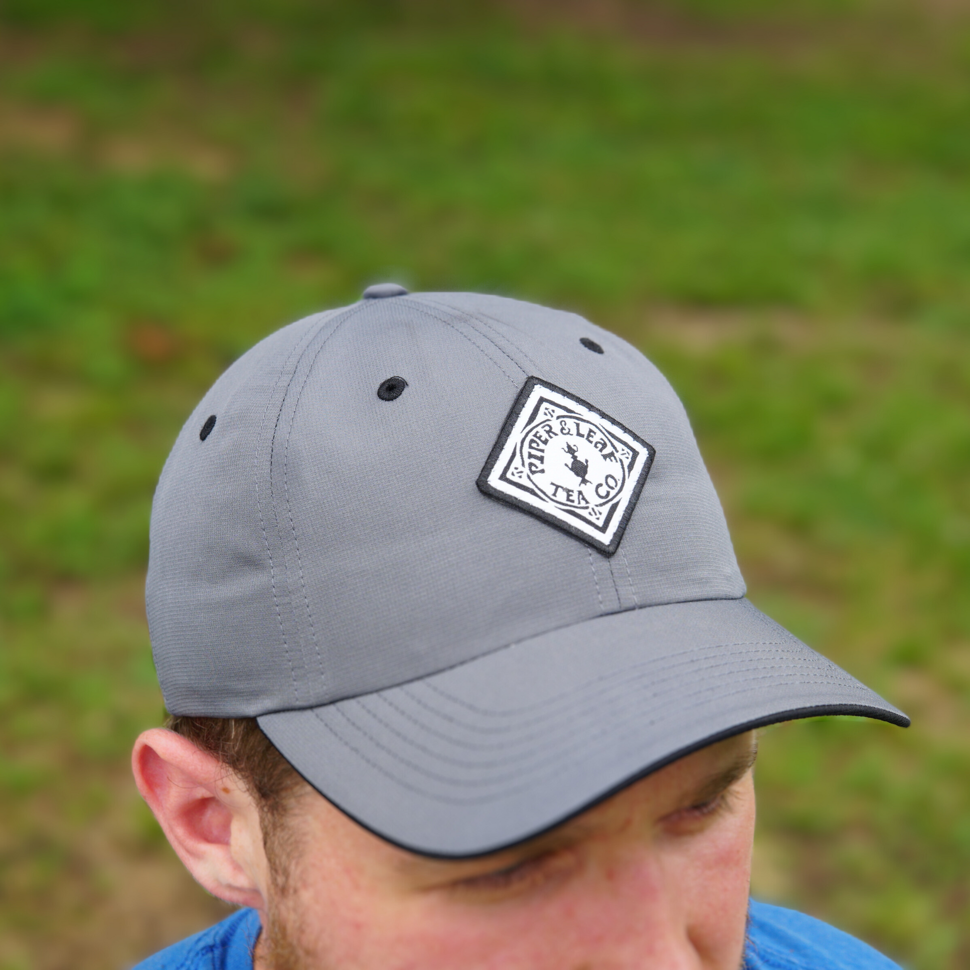 A person wearing a gray Piper & Leaf Tea Co. P&L Sport Cap with a white and black patch on the front, featuring the text "Peerless Team" and a circular emblem, is looking down with a blurred grassy background.