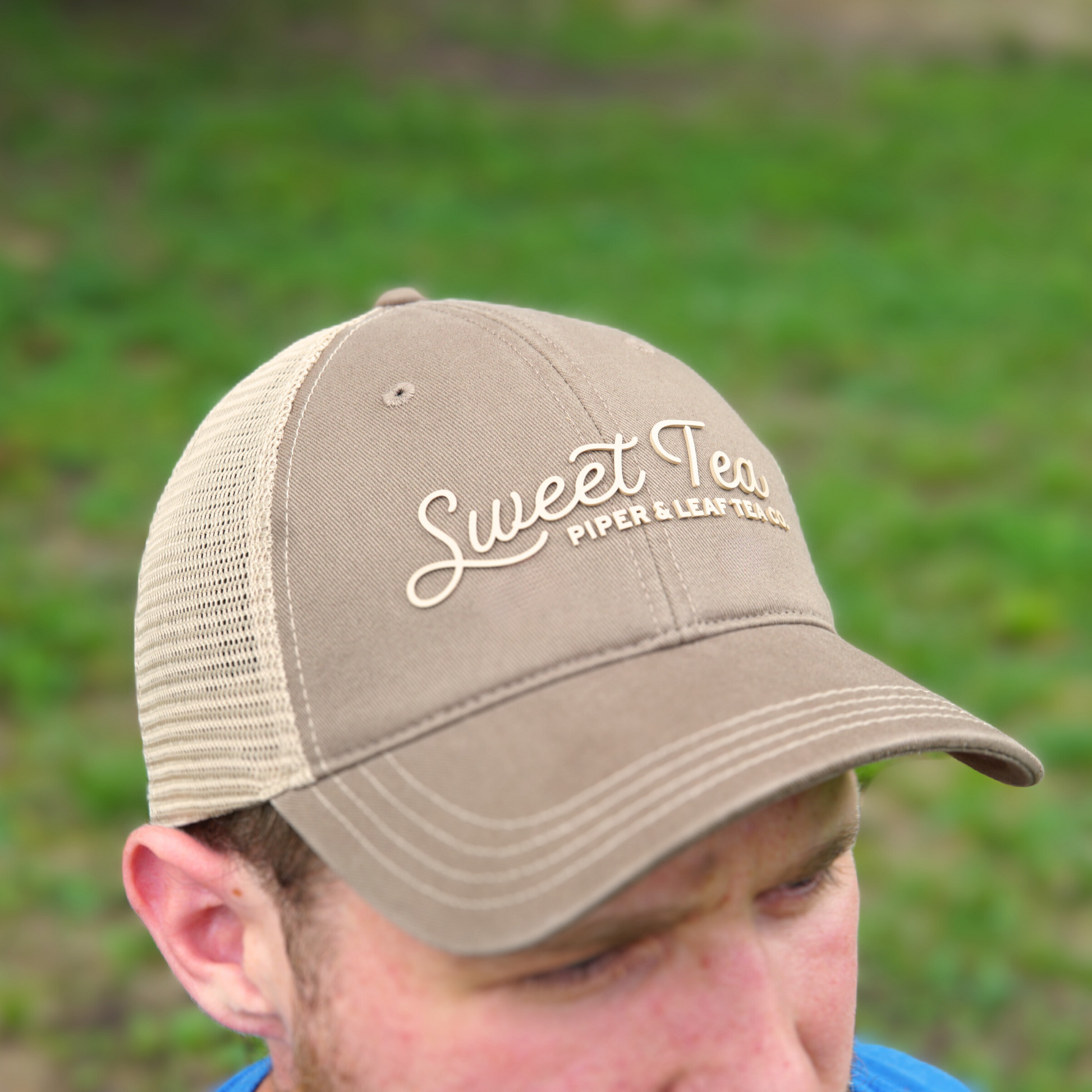 A man wearing a brown P&L Sweet Tea Ball Cap by Piper & Leaf Tea Co. The background features blurred greenery, reminiscent of the South's iconic beverage.