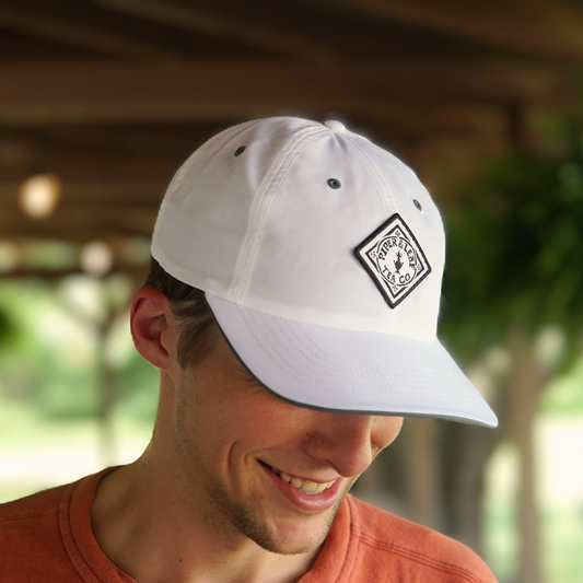 A person wearing an orange shirt and a breathable white P&L Sport Cap from Piper & Leaf Tea Co. with a diamond-shaped logo is smiling and looking down. The background appears to be an outdoor, shaded area with greenery, reminiscent of a Piper & Leaf tea garden.