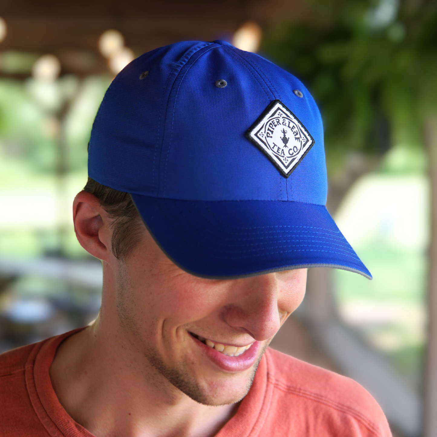 A person wearing a breathable blue P&L Sport Cap with a diamond-shaped Piper & Leaf Tea Co. logo looks down and smiles. The background is blurred with greenery and outdoor scenery.