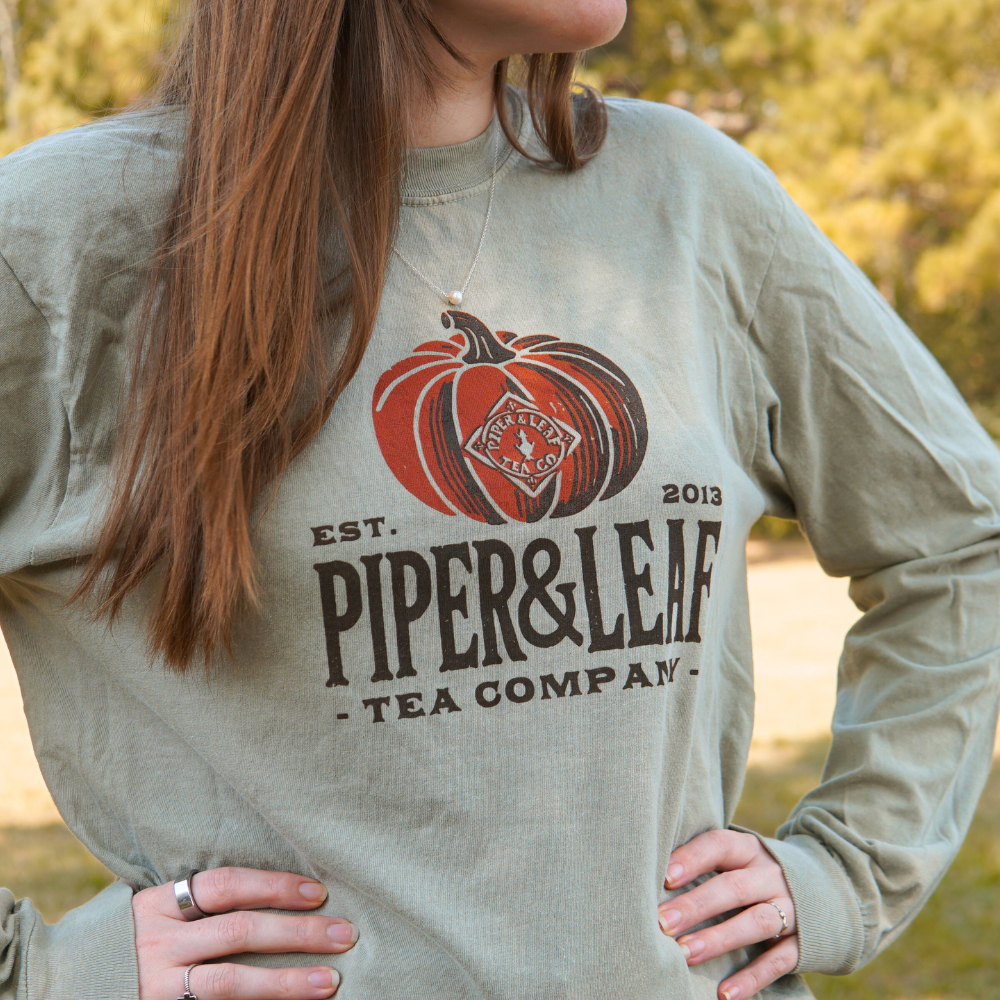 Person wearing the Piper Pumpkin Long Sleeve from Piper & Leaf Tea Co., a sage green shirt with "Piper & Leaf Tea Company Est. 2013" printed alongside an illustration of a Piper pumpkin, perfect for fall festivities.