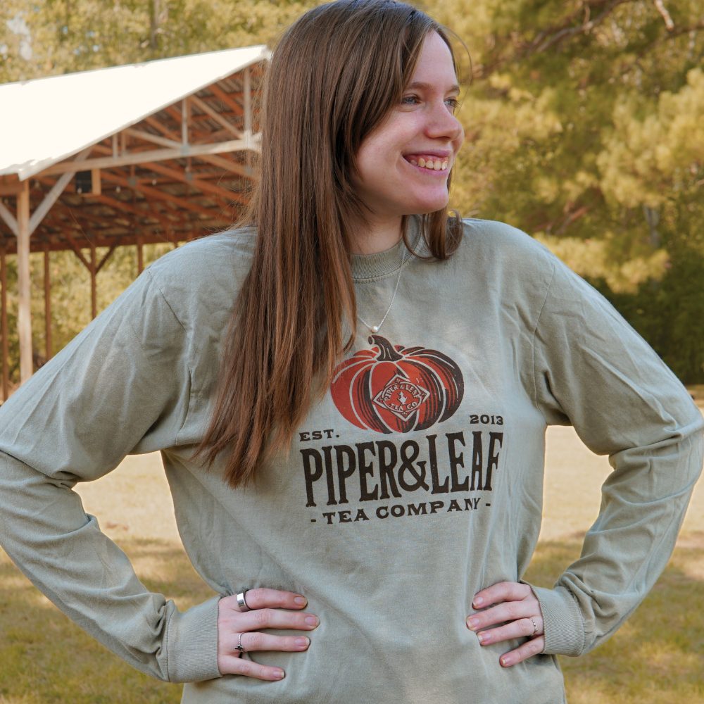 A woman wearing a Piper Pumpkin Long Sleeve shirt from Piper & Leaf Tea Co. stands outdoors, smiling with her hands on her hips. A shelter structure and trees are visible in the background, evoking the spirit of Fall Festivities.