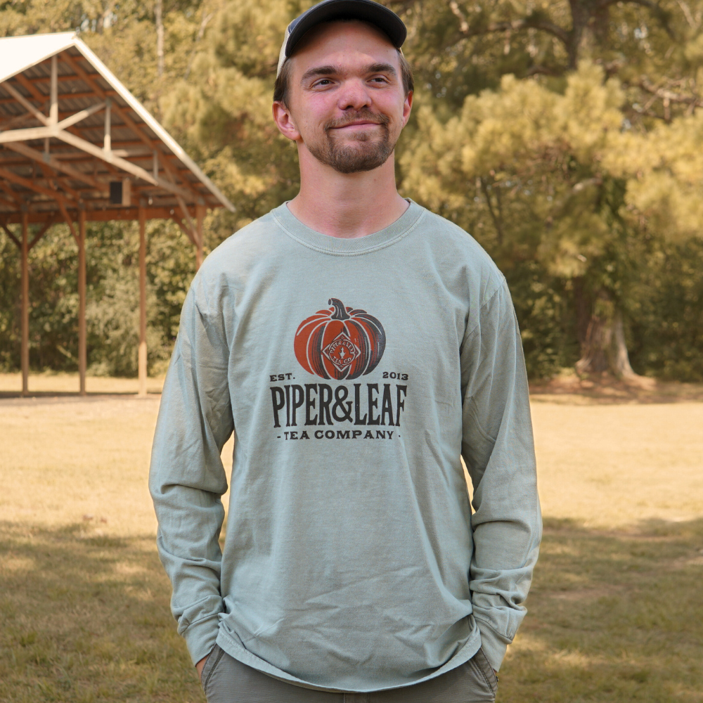 A person wearing a Piper & Leaf Tea Co. Piper Pumpkin Long Sleeve shirt stands outdoors near a wooded area, enjoying the fall festivities.