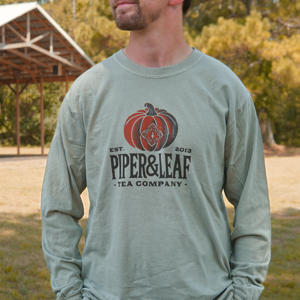 A man wearing the Piper Pumpkin Long Sleeve by Piper & Leaf Tea Co., featuring a sage green color and adorned with "Piper & Leaf Tea Company" alongside a pumpkin graphic, stands outdoors in front of a rustic wooden shed, adding to the charm of fall festivities.