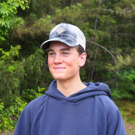 A person in a gray hoodie and the Piper & Leaf Tea Co. Plaid Classic Trucker Hat with an adjustable snapback stands outdoors, smiling, with green trees and foliage in the background.