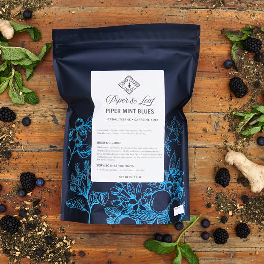 A Piper & Leaf Tea Co. "Piper Mint Blues Pound Bag - 190 servings" is displayed on a wooden surface with fresh mint, blackberries, ginger, and tea leaves around it.