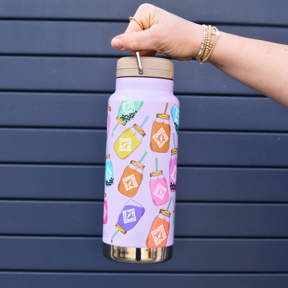 A person holds a purple 32oz Klean Kanteen from Piper & Leaf Tea Co., decorated with colorful honey jar stickers, against a dark striped background. This limited edition bottle features BPA-free stainless steel and double-walled vacuum insulation for optimal beverage temperature.