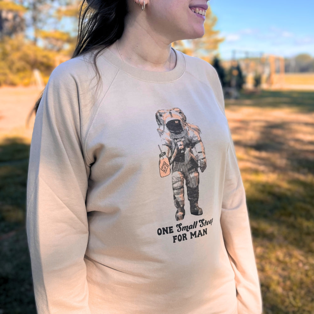 A person wearing the Piper & Leaf Retro Spaceman Sweatshirt by Piper & Leaf Tea Co., featuring a design of a Retro Spaceman and the words "ONE SMALL STEP FOR MAN," stands outside, welcoming the winter's chill.