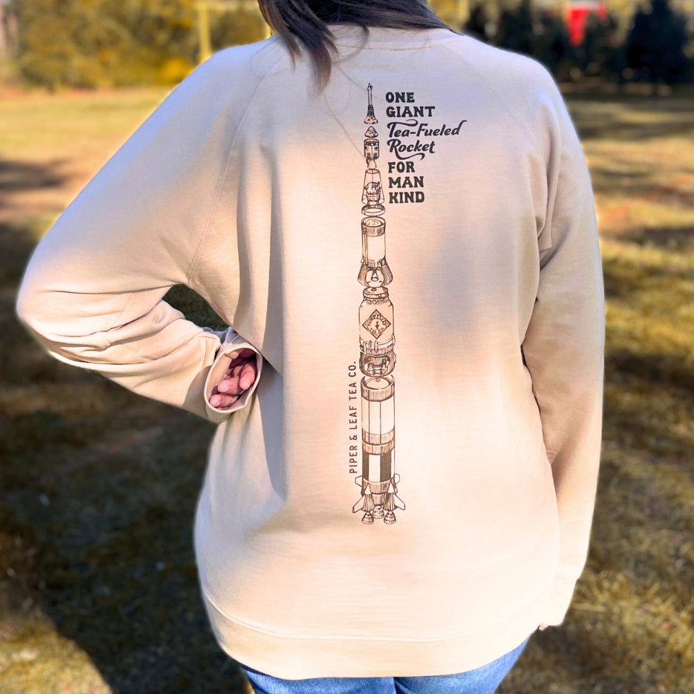 Wrapped in warmth, a person enjoys the comfort of the Piper & Leaf Retro Spaceman Sweatshirt by Piper & Leaf Tea Co. This cozy piece showcases a rocket illustration with the text: "One Giant Tea-Fueled Rocket for Mankind," making it an ideal choice to combat the cold of winter.