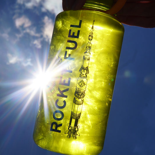 A translucent yellow Piper & Leaf Nalgene Water (Tea) Bottle labeled "Rocket Fuel Tea" held up to the sun, with the silhouette of a rocket depicted on it.