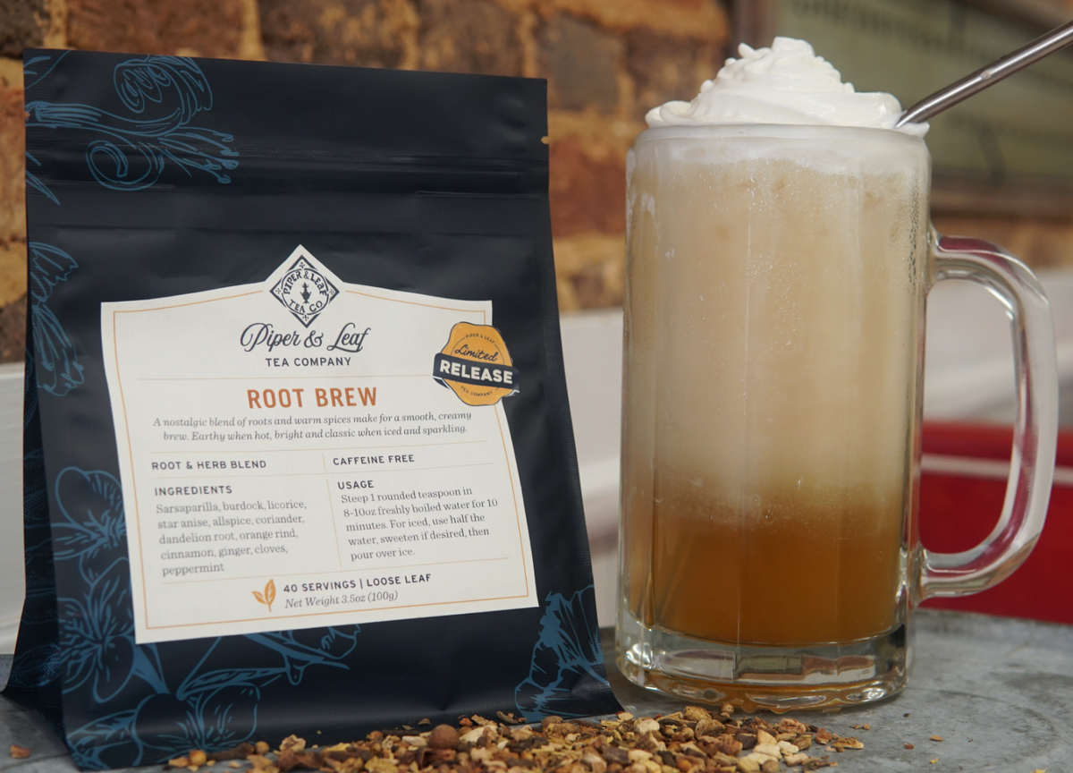 A Root Brew tea bag from Piper and Leaf Tea Co. is beside a glass mug with creamy tea and a metal straw; loose Root Brew leaves and warm spices are scattered in front.