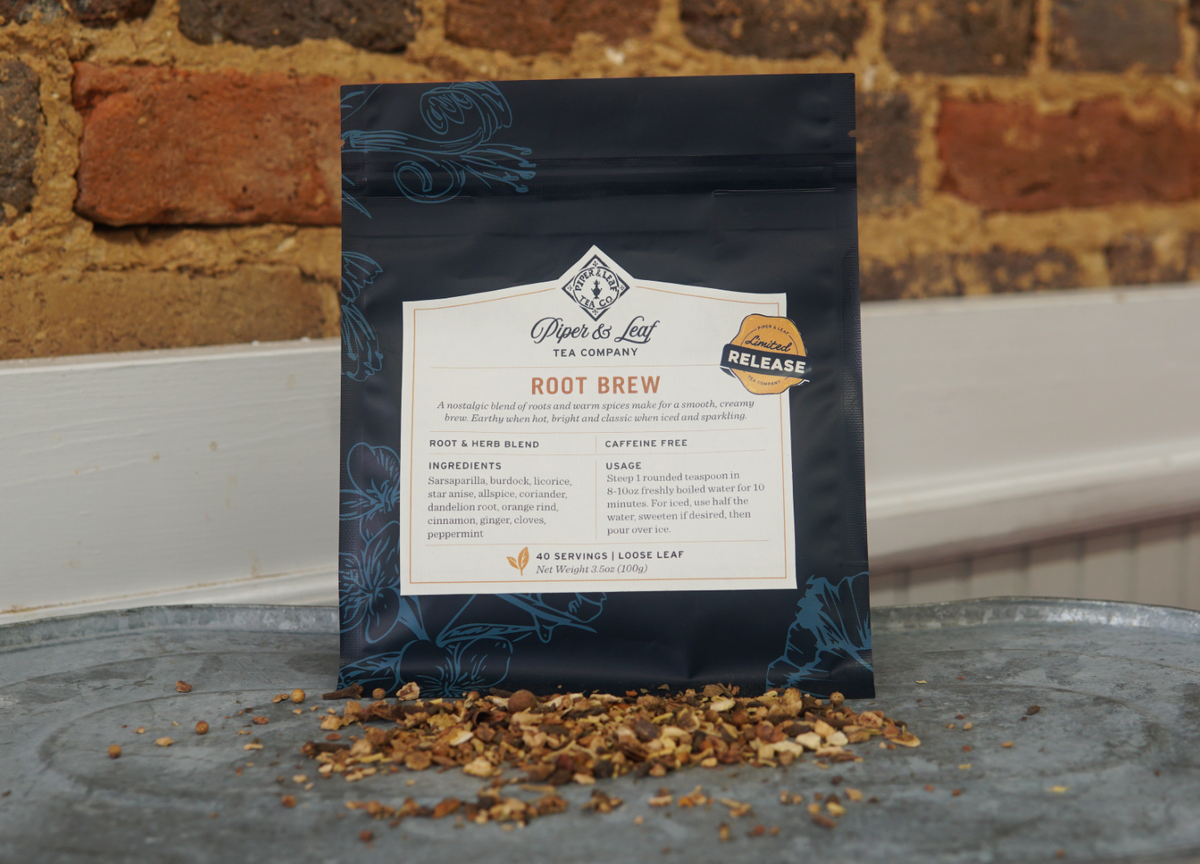 A package of Piper and Leaf Tea Co. Root Brew loose leaf tea rests on a metal surface with warm spices and loose tea scattered in front, set against a brick wall background—a tribute to their classic Root Beer blend.