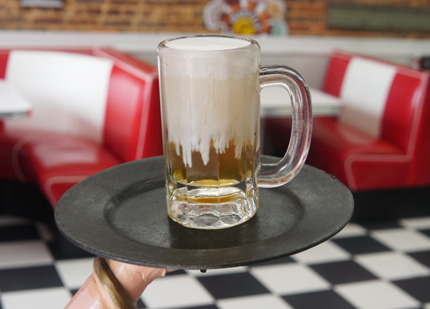 A glass mug of Piper and Leaf Tea Co.'s Root Brew sits on a round tray in a retro diner with red and white booths and a checkered floor.