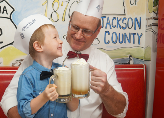 An adult and child, both in paper hats and bow ties, sit on a red booth, smiling and clinking mugs of Piper and Leaf Tea Co.’s Root Brew root beer floats topped with whipped cream.