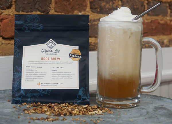A bag of Piper and Leaf Tea Co. Root Brew loose leaf tea rests beside a glass of iced tea topped with whipped cream and a metal straw, set on a table in front of a brick wall.