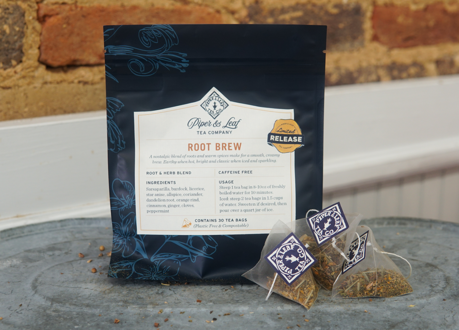 A bag of Piper and Leaf Tea Co. Root Brew and three pyramid tea bags rest on a metal surface; the creamy, spiced blend is displayed in front of a rustic brick wall.