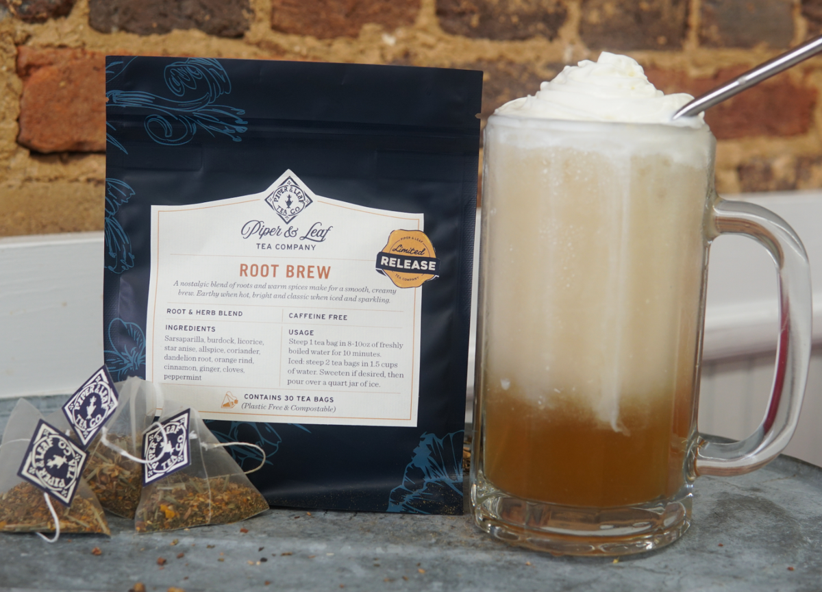 A glass mug of creamy Root Brew tea topped with whipped cream sits beside a Piper and Leaf Tea Co. Root Brew blend bag and two pyramid tea bags on a wooden surface.