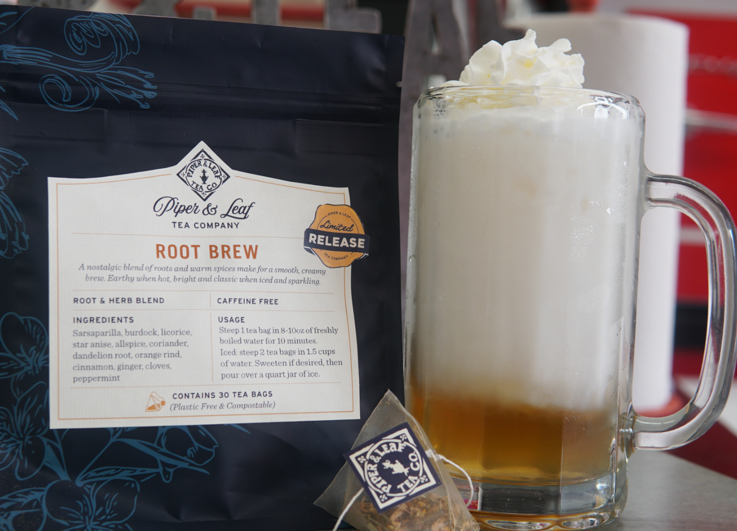 A glass mug of creamy brew topped with whipped cream sits next to a Piper and Leaf Tea Co. Root Brew tea bag and its packaging, which features nostalgic spices along with ingredients and usage instructions.
