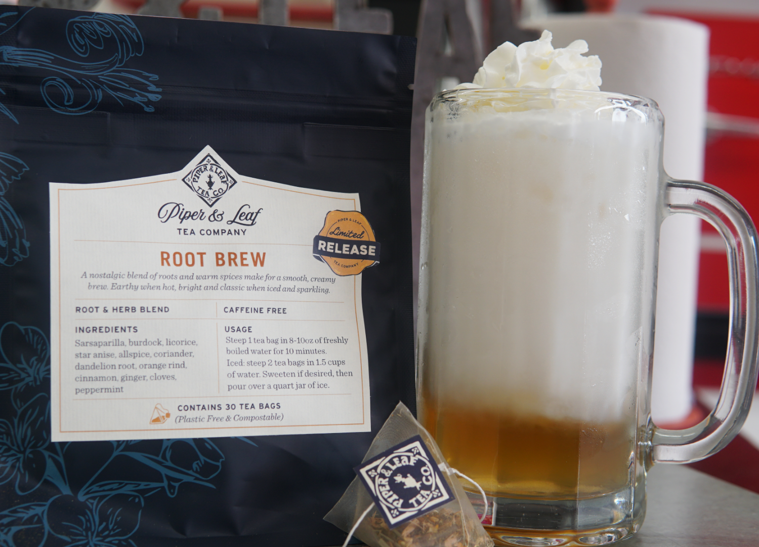 A glass mug of creamy brew topped with whipped cream sits next to a Piper and Leaf Tea Co. Root Brew tea bag and its packaging, which features nostalgic spices along with ingredients and usage instructions.