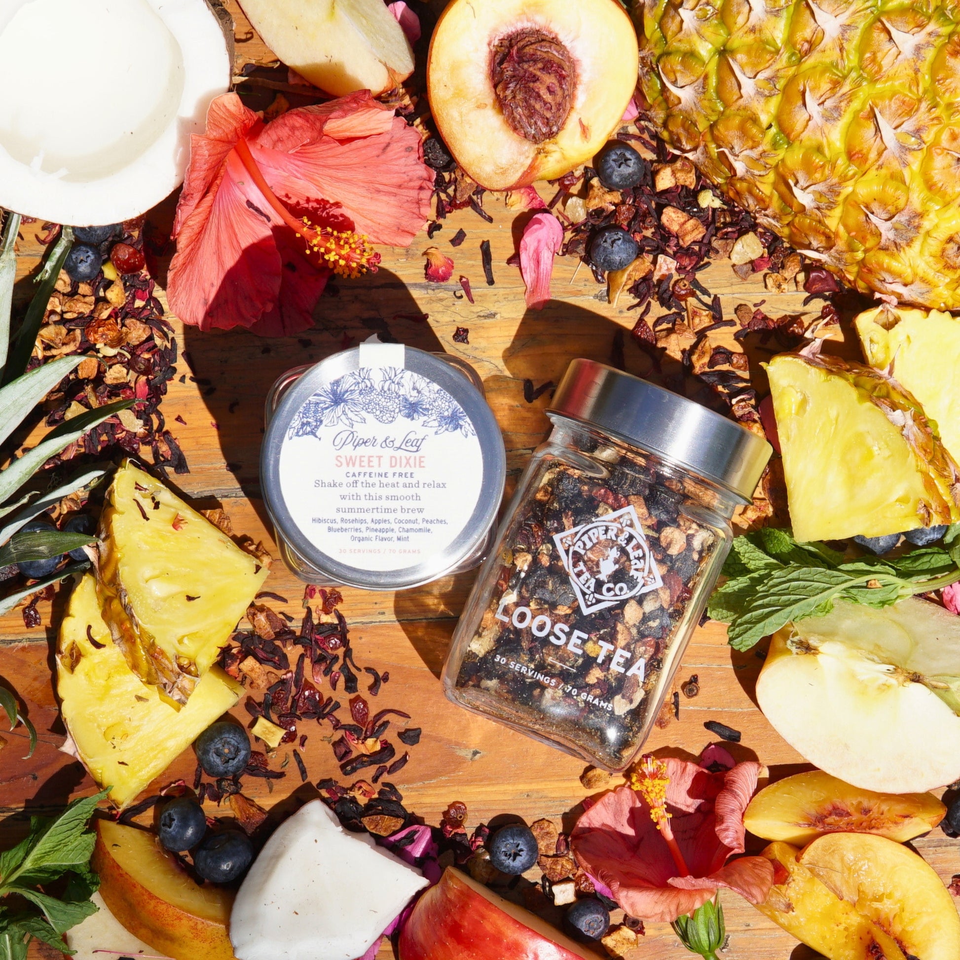 A Piper & Leaf Tea Co. Sweet Dixie Glass Jar of Loose Leaf Tea is surrounded by pineapple, apple, and peach slices, with scattered hibiscus flowers and blueberries nearby.