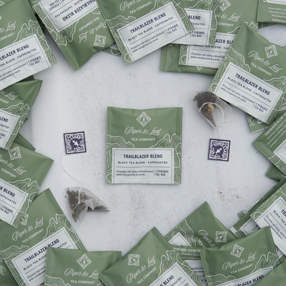 Various packets of Piper & Leaf Tea Co.'s Trailblazer Blend Case of Individually Wrapped Tea Bags - 50ct in green packaging are scattered on a white surface, with some individually packaged tea sachets visible outside the packets.