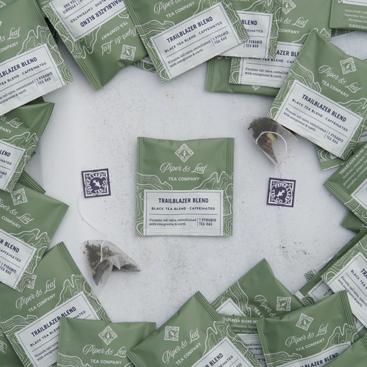 Various packets of Piper & Leaf Tea Co.'s Trailblazer Blend Case of Individually Wrapped Tea Bags - 50ct in green packaging are scattered on a white surface, with some individually packaged tea sachets visible outside the packets.