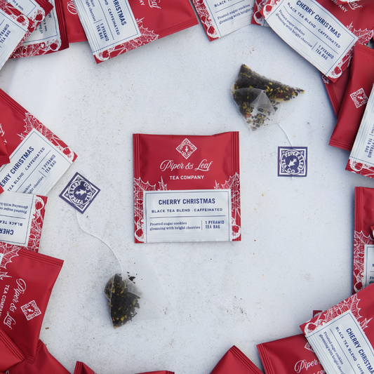 A festive flat lay displays Piper & Leaf Tea Co.'s "Cherry Christmas" case of individually wrapped red tea bags (50ct), making it perfect for gift baskets. The charming scene is enhanced by a few pyramid-shaped, individually packaged tea sachets.