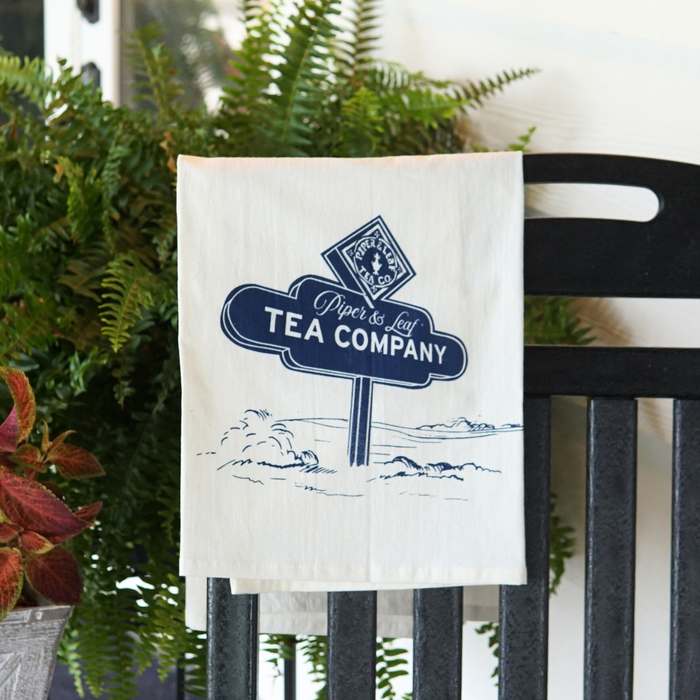 A white tea towel hangs on a black chair, featuring a blue vintage-style sign with the text "Piper & Leaf Tea Company" amidst illustrated clouds and leaves. This charming piece captures the essence of Lacey's Spring. This is the Tea Towel-P&L Road Sign from Piper & Leaf Tea Co.