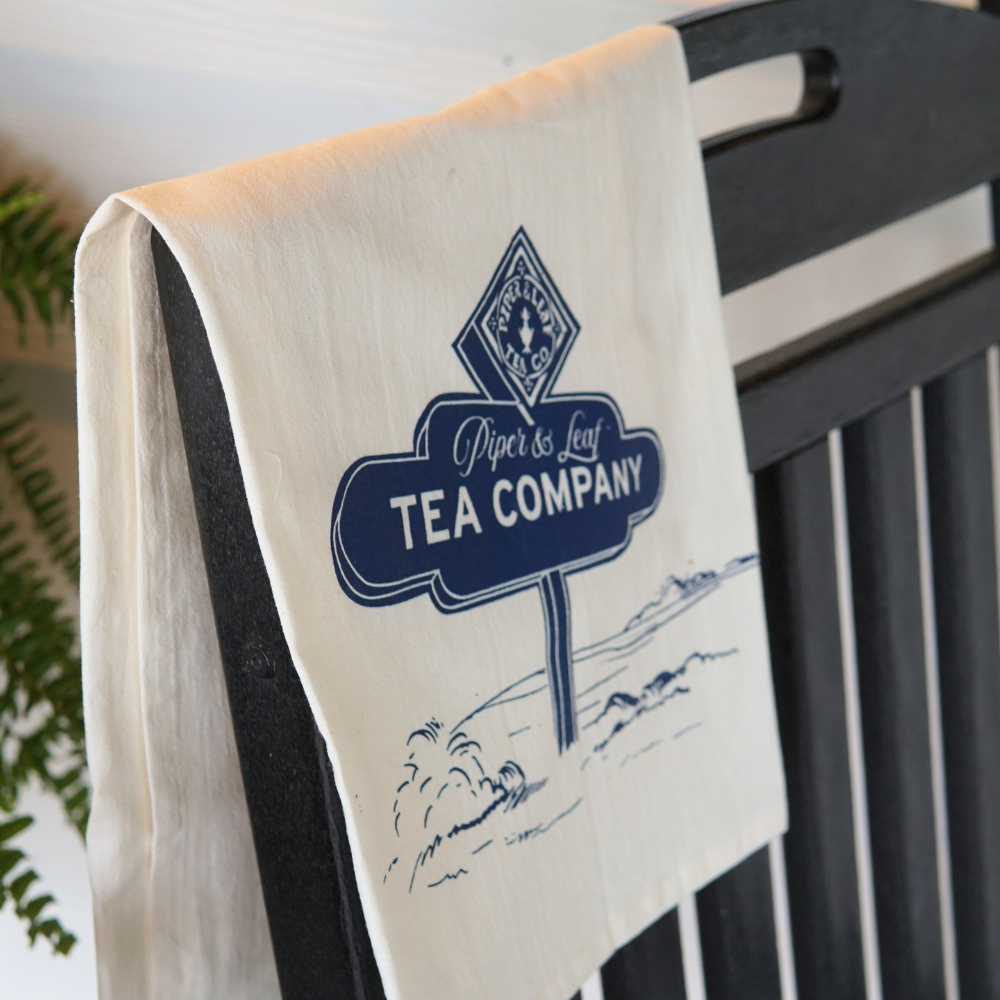 A white Tea Towel-P&L Road Sign with "Piper & Leaf Tea Co." inscribed on it hangs over the back of a black wooden chair.