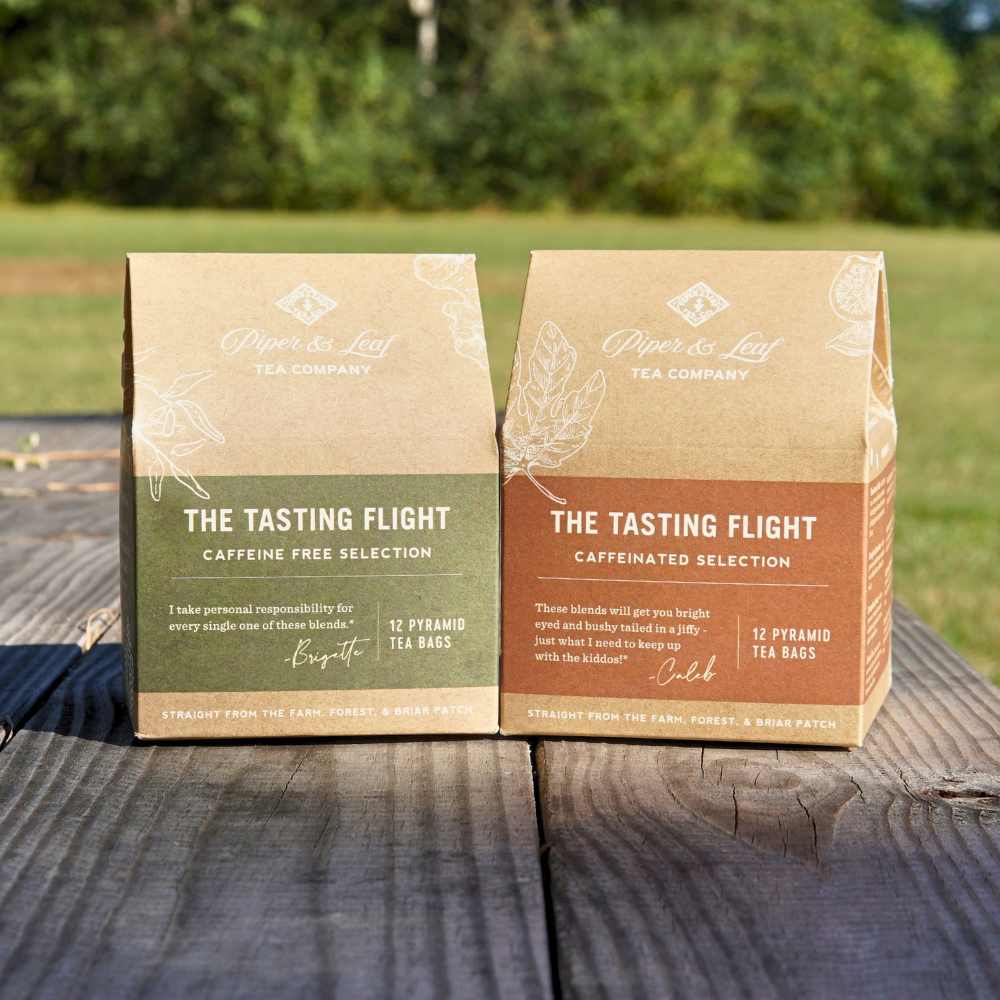 Two boxes of Piper & Leaf Tea Co. are placed on a wooden table amidst the greenery. The green-labeled "Caffeine Free Selection" and the brown "Caffeinated Selection" from the Tasting Flight Duo offer an assortment of unique blends to enjoy throughout the year.