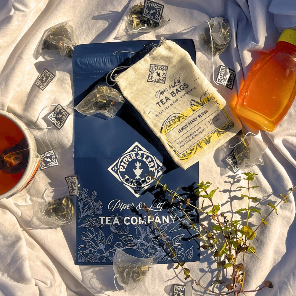 Piper & Leaf Tea Co.'s Tea Subscription blends and packaging are beautifully displayed on a fabric background, featuring a tea cup and honey bottle. Scattered plants enhance the rich artisan tea experience.