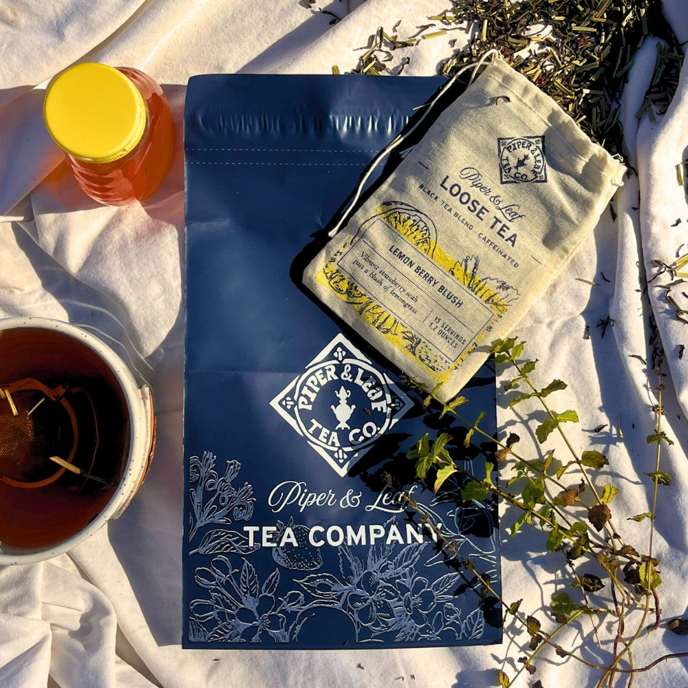 Discover the artisan tea experience with our promotional setup featuring a blue bag, Piper & Leaf Tea Co.'s lemon mint blend, a honey jar, and a loose tea leaves cup on white fabric. Indulge in the finest Tea Subscription selections today.