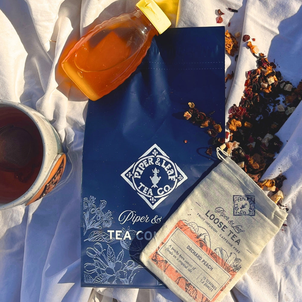 A blue tea bag, a pouch labeled "Orchard Peach Loose Tea," and an artisan tea mug from Piper & Leaf Tea Co., offered through their Tea Subscription, sit alongside a bottle of honey on a white cloth.